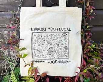 Puede incluir: Bolso tote de color crema con estampado negro. El diseño presenta varias ilustraciones de ranas y setas, con el texto "SUPPORT YOUR LOCAL FROGS". El bolso tiene dos asas y está hecho de una tela resistente.