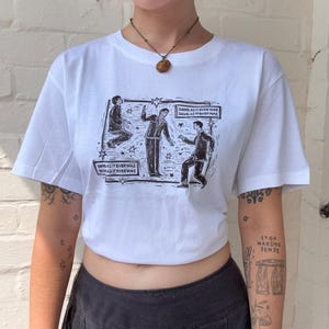 May include: A white short-sleeved t-shirt featuring a black graphic print of three figures in suits. The design includes stars and two rectangular speech bubbles containing the repeated text, "SAME AS IT EVER WAS SAME AS IT EVER WAS."