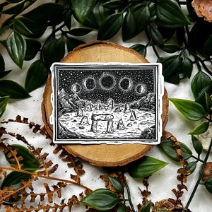 May include: Black and white sticker featuring a landscape with mountains, standing stones, and phases of the moon. The sticker is rectangular with a white border and is placed on a wooden slice surrounded by greenery.