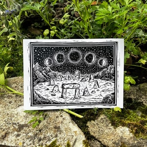 May include: A black and white linocut print depicting a celestial scene with phases of the moon, a starry sky, and a stone circle. The print is set against a natural background of green foliage and stone.