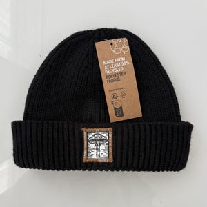 May include: A black ribbed knit beanie featuring a rectangular patch with a white mushroom illustration on a brown border. A brown cardboard tag is attached, printed with the text: MADE FROM AT LEAST 50% RECYCLED POLYESTER FABRIC.