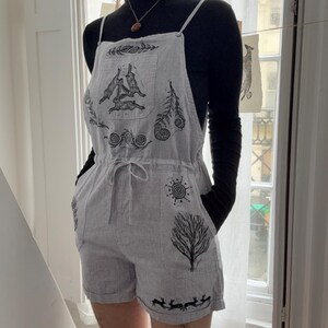 Könnte beinhalten: Ein hellgrauer Cord-Overall mit dünnen Trägern, Kordelzug in der Taille und schwarzen Illustrationen von drei Hasen, Farnen, einer Sonne und einem Baum, getragen über einem schwarzen Langarmshirt mit einer schlichten Halskette.