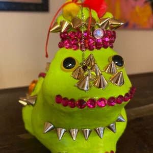 Peut inclure: Une figurine de grenouille en céramique verte avec des pointes dorées et argentées, des strass roses et un pompon rouge et blanc sur la tête.