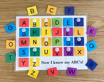 May include: A colorful alphabet learning chart with letter tiles in a grid pattern. The chart has the letters A through Z in order, with each letter on a separate tile. The tiles are arranged in a grid pattern, with each row containing 7 tiles. The tiles are all different colors, and the letters are printed in black. The chart is on a wooden surface. The text "Now I know my ABC's!" is printed at the bottom of the chart.