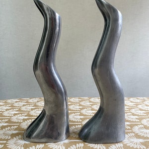 May include: Two silver, abstract, sculptural vases with a wavy, organic design. The vases are made of metal and have a smooth, polished finish.