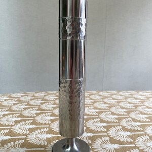 Può includere: Un vaso in metallo argentato martellato con una base rotonda. Il vaso ha una finitura liscia e lucida e una texture martellata nella sezione centrale.