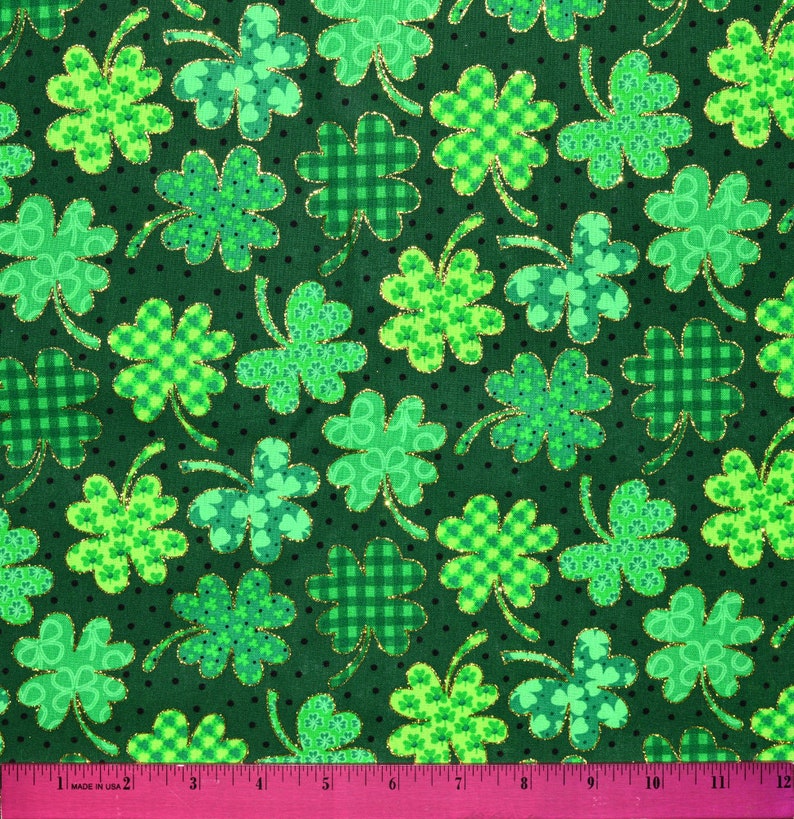 Op de afbeelding: Groene stof met een herhalend patroon van vierbladige klavers. De klavers zijn gemaakt van verschillende tinten groen en hebben een gouden glitterrand. De achtergrond is donkergroen met kleine witte stippen.