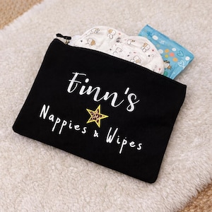 May include: A black zipped pouch with the text "Finn's Nappies & Wipes" in white script, featuring a gold star with a leopard print centre. The pouch contains baby items, including a white cloth with bee patterns and a blue packet.