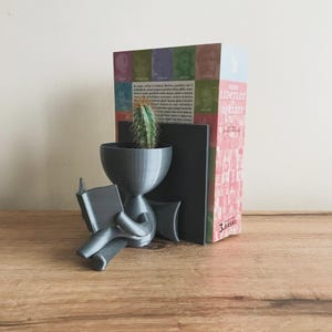May include: A gray 3D printed bookend shaped like a person sitting and reading a book. The bookend has a pot on its head with a small cactus plant. The bookend is holding a book in its left hand and is sitting on a wooden surface. The bookend is holding up a book with the title "Tutku Liderler ve Aşıkları" on the spine. The book is in Turkish.