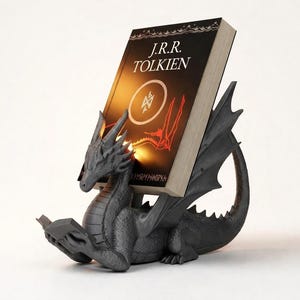 Puede incluir: Un sujetalibros de dragón gris oscuro que sostiene un libro de J.R.R. Tolkien. El dragón está esculpido con detalles intrincados, incluyendo alas y una cola. El libro tiene una cubierta negra con texto blanco y un diseño circular.