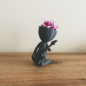 May include: A gray 3D printed planter shaped like a stick figure playing a guitar. The planter has a flower with pink petals in the head.