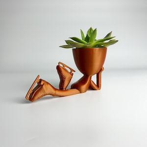 Könnte beinhalten: Ein kupferfarbener, 3D-gedruckter Pflanztopf in Form einer liegenden Person mit hochgestreckten Beinen, die Schlittschuhe trägt. Der Topf hat einen runden Kopf und eine Sukkulente wächst darin.