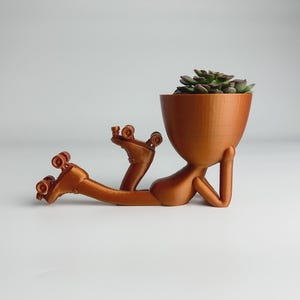 Op de afbeelding: Een koperkleurige plantenpot in de vorm van een persoon op rol schaatsen, met een vetplant in het hoofd van de pot.