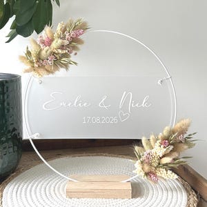 Peut inclure: Panneau de mariage en acrylique transparent avec les noms "Emelie & Nich" et la date "17.08.2026" en écriture blanche. Décoré de compositions florales séchées roses, beiges et marron. Il repose sur un socle en bois.