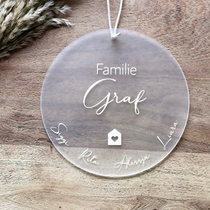 Könnte beinhalten: Ein rundes Schild aus klarem Acryl mit dem Text "Familie Graf" und den Namen "Siggi", "Rita", "Alessia" und "Liara", die um ein Haussymbol mit einem Herzen darin geschrieben sind.