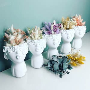 Op de afbeelding: Vijf witte keramische potten in de vorm van gezichten met gedroogde bloemenarrangementen. De potten hebben bloemdetails rond het hoofd en een gesloten-lip pruilmond. De bloemen zijn in tinten roze, paars, wit en geel.
