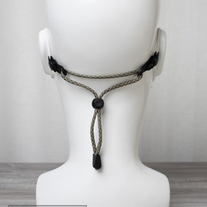 May include: A close-up of a black adjustable strap with a green and brown camouflage pattern. The strap is attached to a white mannequin head. The strap has a black toggle for adjusting the length.