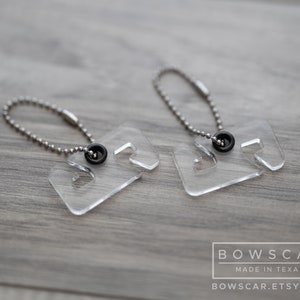May include: Two clear acrylic keychains with a square shape and a cutout design. Each keychain has a small black circular accent and a silver ball chain. The keychains are displayed on a gray wooden surface.