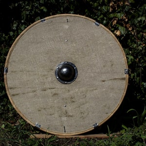 Op de afbeelding: Een rond, houten schild met een metalen boss in het midden. Het schild is bedekt met een beige stof en heeft metalen accenten rond de rand.