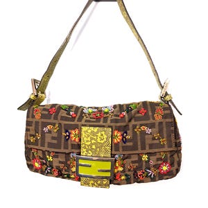 Może przedstawiać: Brązowa torebka z powtarzającym się wzorem logo, ozdobiona kolorowymi, koralikowymi wzorami kwiatowymi. Torebka posiada żółto-zielone zapięcie i zielony pasek. Torebka zawieszona na białym tle.