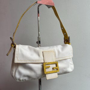 May include: White handbag with a yellow leather strap and a gold-coloured buckle. The bag has a rectangular shape with a flap closure. The stitching is yellow, and the bag is displayed on a metal stand.