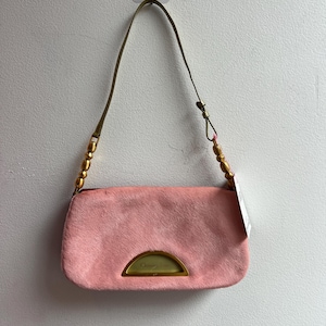 Puede incluir: Un bolso rectangular rosa con asa y detalles dorados. El bolso tiene una superficie texturizada y un detalle semicircular dorado en la parte delantera. El asa es verde oliva con cuentas doradas.
