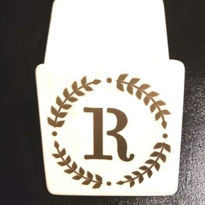 May include: White ceramic holder with a gold laurel wreath design and the letter "R" in the center.