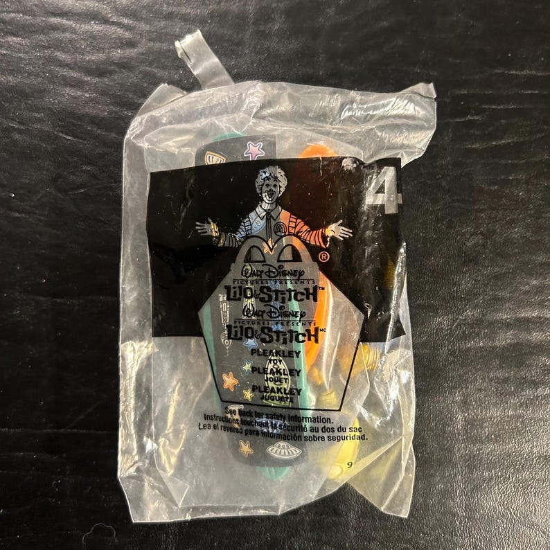 May include: Sealed McDonald's Happy Meal toy featuring a Pleakley character from Disney's Lilo & Stitch. The clear plastic bag displays the movie logo, character name, and the number 4. The toy is colorful with green, orange, and yellow accents.
