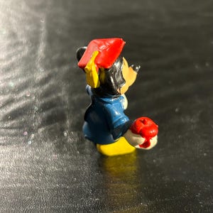 Puede incluir: Una peque&ntilde;a figura de pl&aacute;stico de Mickey Mouse con un gorro de graduaci&oacute;n rojo y un atuendo azul y amarillo. Est&aacute; sosteniendo una manzana roja en su mano derecha.