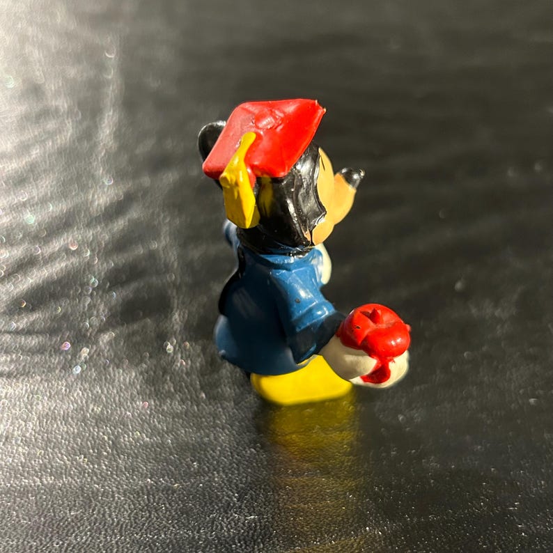 Puede incluir: Una peque&ntilde;a figura de pl&aacute;stico de Mickey Mouse con un gorro de graduaci&oacute;n rojo y un atuendo azul y amarillo. Est&aacute; sosteniendo una manzana roja en su mano derecha.