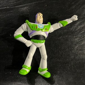 May include: A Buzz Lightyear action figure in a dynamic pose. The figure is mainly white with green and purple accents, including a green chest plate and accents on the arms and legs. The figure has a smiling expression and is posed with one arm raised.