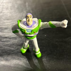 May include: A small Buzz Lightyear action figure in a dynamic pose. The figure is mainly white with green and purple accents. It has a smiling face and is posed with one arm extended and the other bent at the elbow.