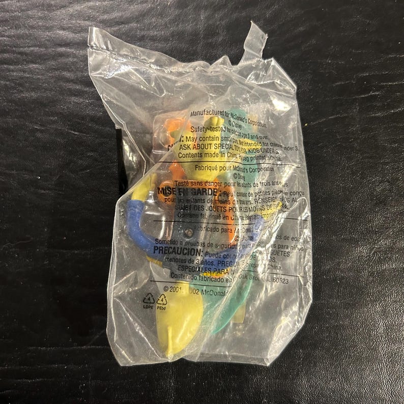 May include: A sealed plastic bag containing a colorful toy. The toy appears to be a small, multi-colored figure with yellow, blue, green, and orange elements. The packaging includes printed text and safety warnings.