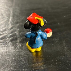 Puede incluir: Una peque&ntilde;a figura de pl&aacute;stico de Mickey Mouse, negra y azul, que lleva una gorra de graduaci&oacute;n roja y sostiene una manzana. La figura tiene un borla amarilla en la gorra.