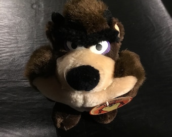 May include: Brown plush toy of Taz the Tasmanian Devil from the Looney Tunes cartoon. The toy has a purple eye and a black nose. The toy is holding its arms in front of it. The toy has a tag that says "Warner Bros."