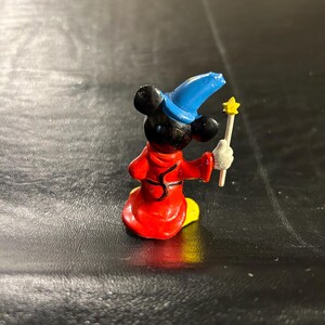 May include: A small figurine of Mickey Mouse in a red robe and blue wizard hat, holding a yellow star-tipped wand. The figure has black ears and a yellow base. The figurine is a collectible item.