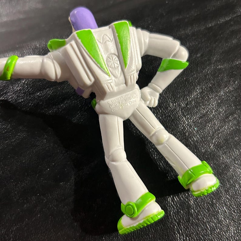 May include: A white Buzz Lightyear action figure with green and purple detailing. The figure has a white body with green trim on the arms, legs, and feet. The head is purple, and the figure is posed with arms outstretched.