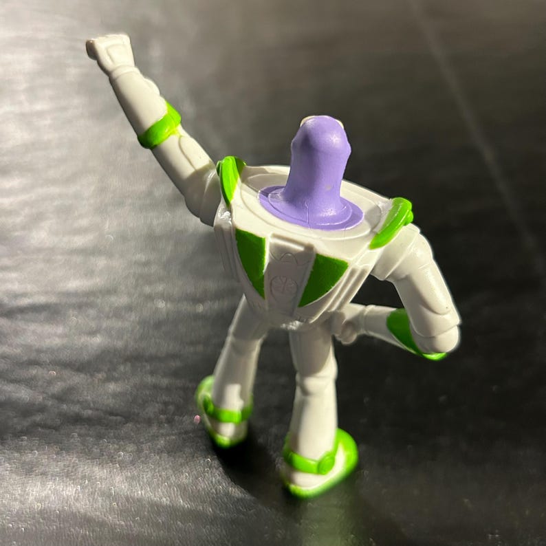 May include: A white and green Buzz Lightyear action figure with a purple head. The toy is in a dynamic pose, with one arm raised. The figure has green accents on the arms, legs, and feet. The toy is a collectible item.