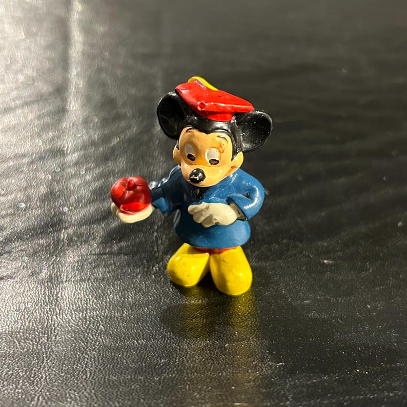 Puede incluir: Una peque&ntilde;a figura de pl&aacute;stico de Mickey Mouse con una t&uacute;nica de graduaci&oacute;n azul y amarilla y un gorro de graduaci&oacute;n rojo. Est&aacute; sosteniendo una manzana roja en su mano izquierda.