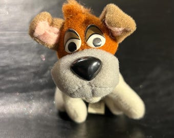 May include: A plush toy dog with brown and white fur, large black eyes, and a black nose. The dog is sitting with its head tilted slightly to the side.