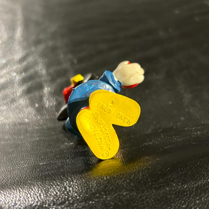 Puede incluir: Un zapato de juguete de pl&aacute;stico amarillo con el texto "DISNEY" y "HONG KONG" impreso en &eacute;l. El zapato es parte de una figura de juguete m&aacute;s grande.