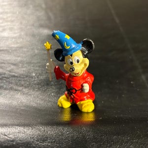 May include: A small figurine of Mickey Mouse dressed as a sorcerer. Mickey wears a red robe, yellow shoes, and a blue hat with gold stars and a crescent moon. He holds a yellow star-tipped wand.