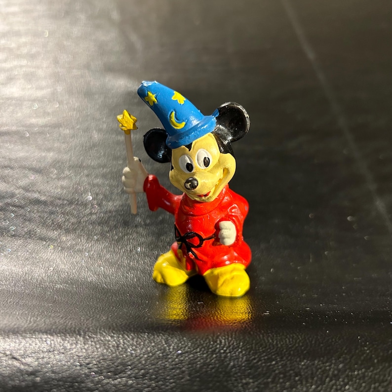May include: A small figurine of Mickey Mouse dressed as a sorcerer. Mickey wears a red robe, yellow shoes, and a blue hat with gold stars and a crescent moon. He holds a yellow star-tipped wand.
