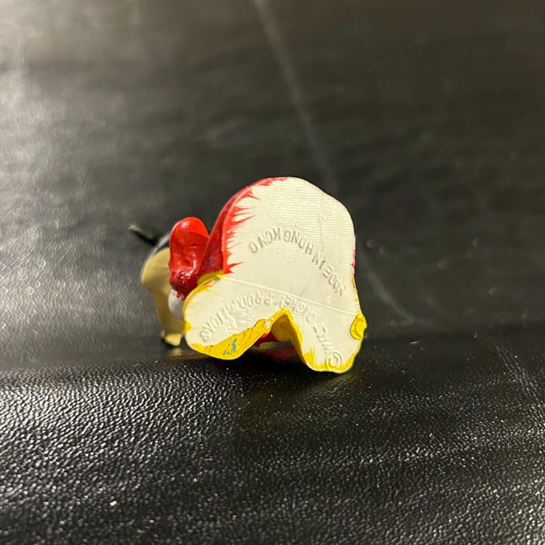 May include: A small, colorful figurine of a cartoon character, viewed from the back. The figurine has a red hat, yellow and white clothing, and black ears. The base of the figurine has the text "MADE IN HONG KONG" and "&copy; DISNEY PRODUCTIONS".