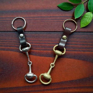 May include: Two keyrings featuring horse bit designs. One has a black leather strap and a silver-coloured bit, the other has a brown leather strap and a brass-coloured bit. Both have a metal ring.