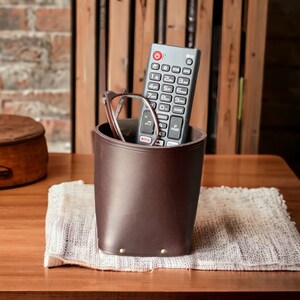 May include: A brown leather remote control holder with a silver remote control and glasses inside. The holder is sitting on a white crocheted placemat.