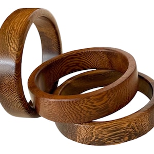 Puede incluir: Tres brazaletes de madera con un patrón de veta de madera marrón. Los brazaletes están apilados uno encima del otro.