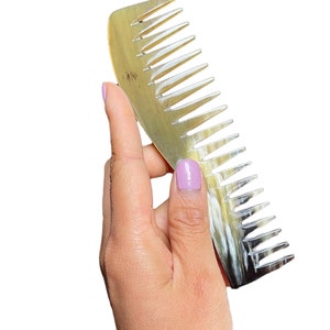 Pode incluir: Uma mão segura um pente de chifre marrom e preto. O pente tem uma forma curva e mede aproximadamente 25 cm de comprimento.
