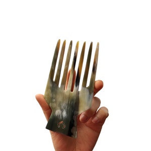 Puede incluir: Una mano sostiene un peine marrón y negro con un diseño único. El peine tiene una base ancha y varios dientes.