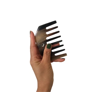 May include: A brown and black comb made of horn with seven teeth. The comb is held in a hand with green nail polish.